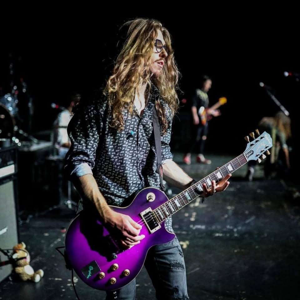 A man with long, curly hair plays a purple electric guitar on stage.