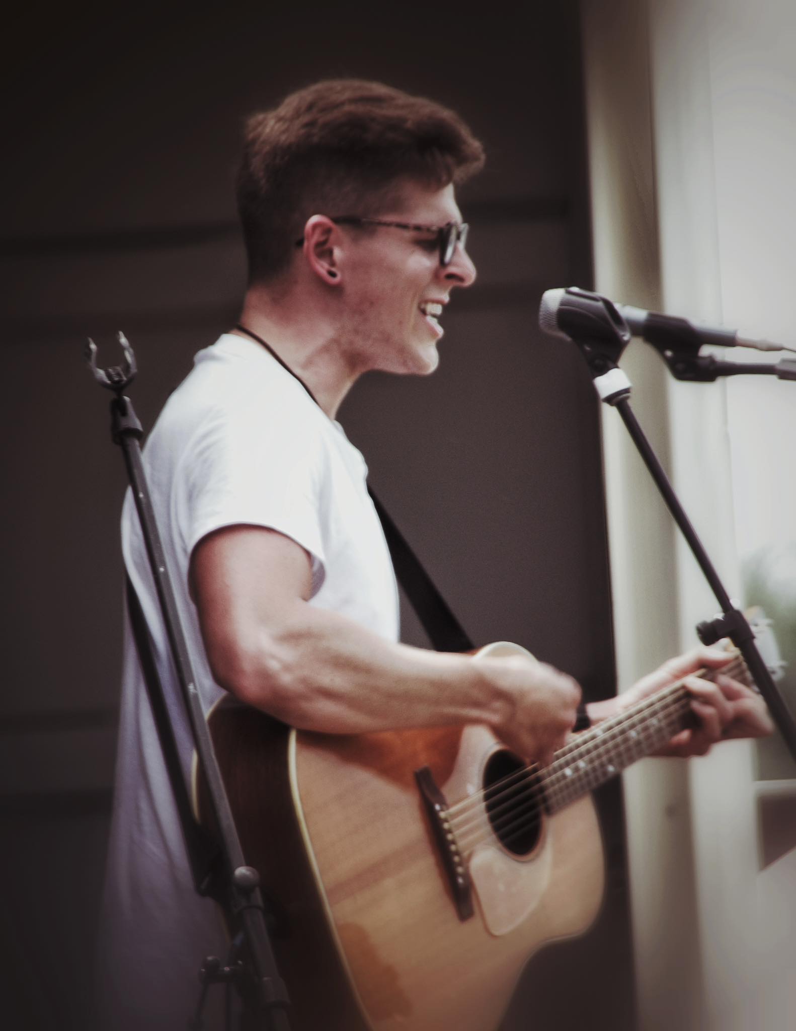 Young man with glasses playing acoustic guitar and singing into a microphone, smiling.