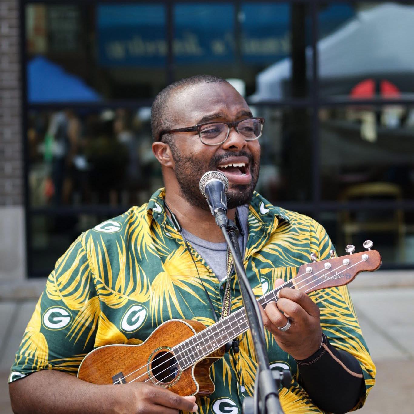 Man in green & yellow Hawaiian shirt plays ukulele & sings into microphone.