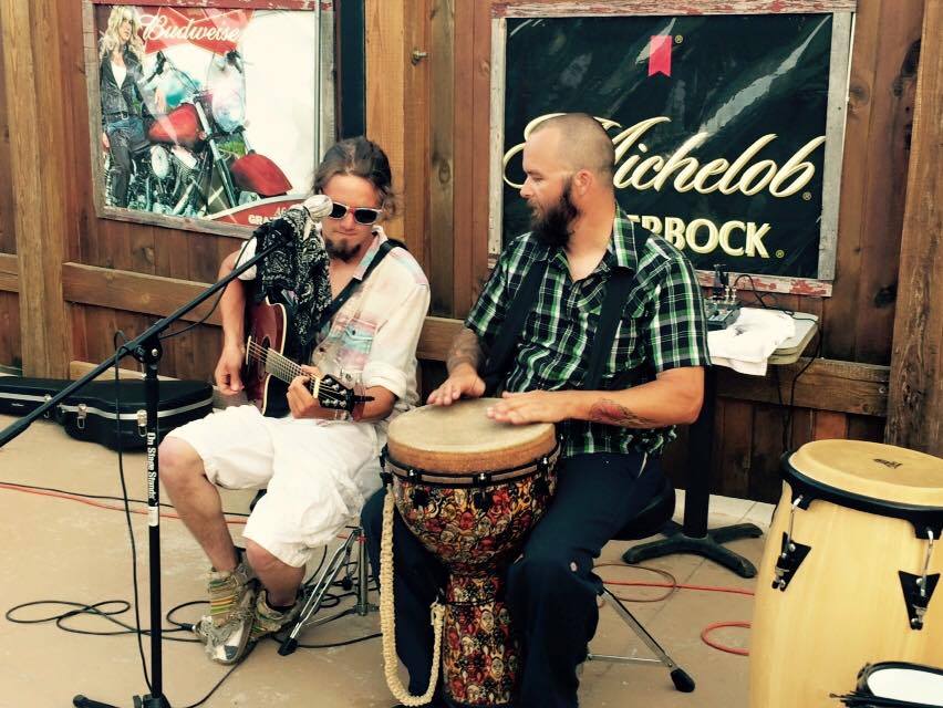 Two men play music outside a bar. One plays guitar, the other plays drums.