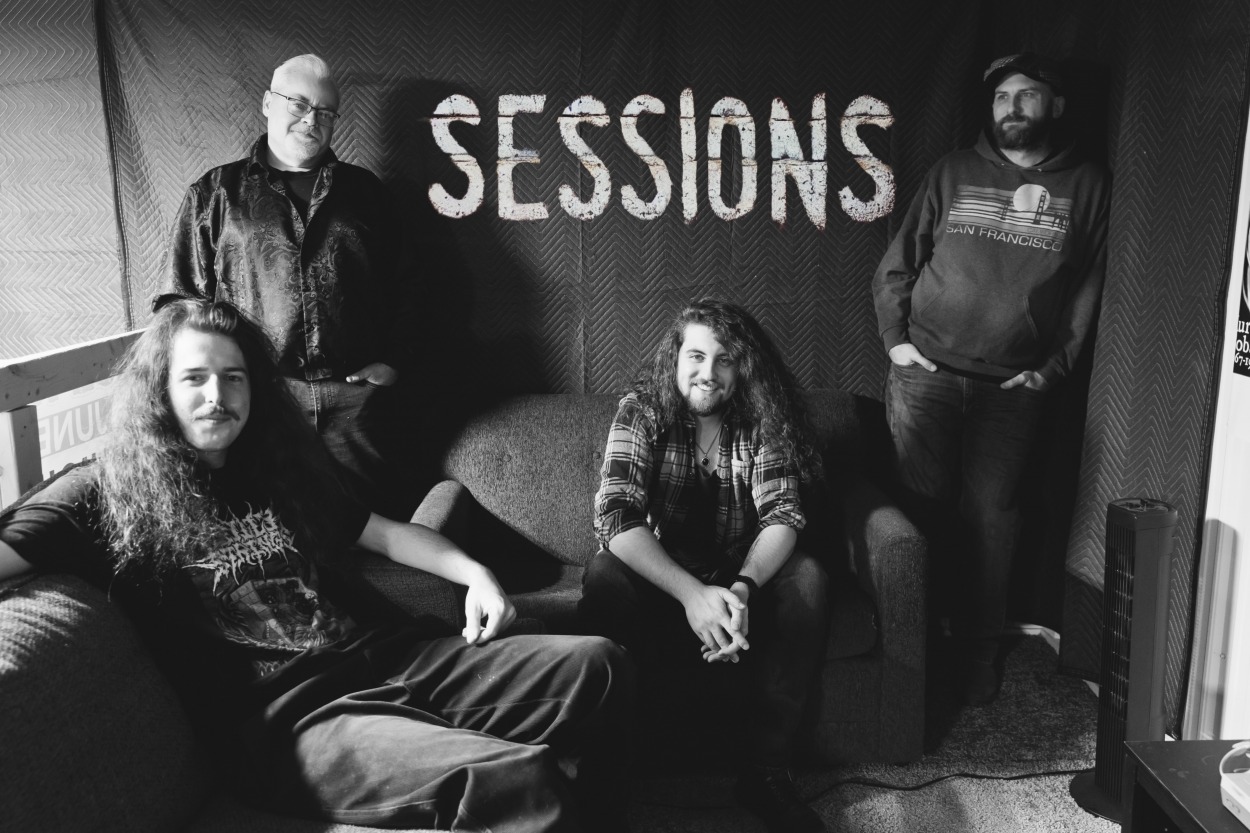 Four men pose in front of a wall with SESSIONS painted on it.