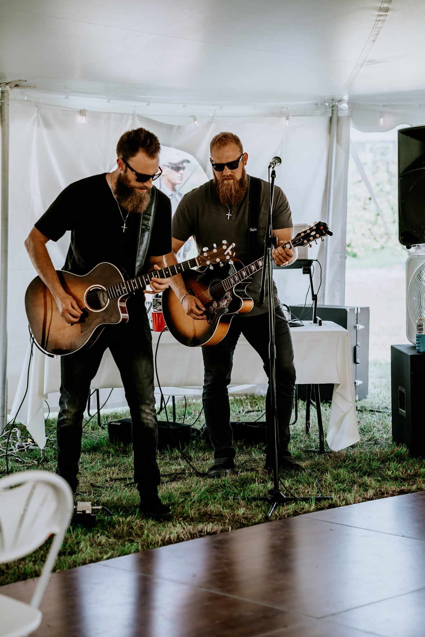 Two men with beards and sunglasses play acoustic guitars under a tent.
