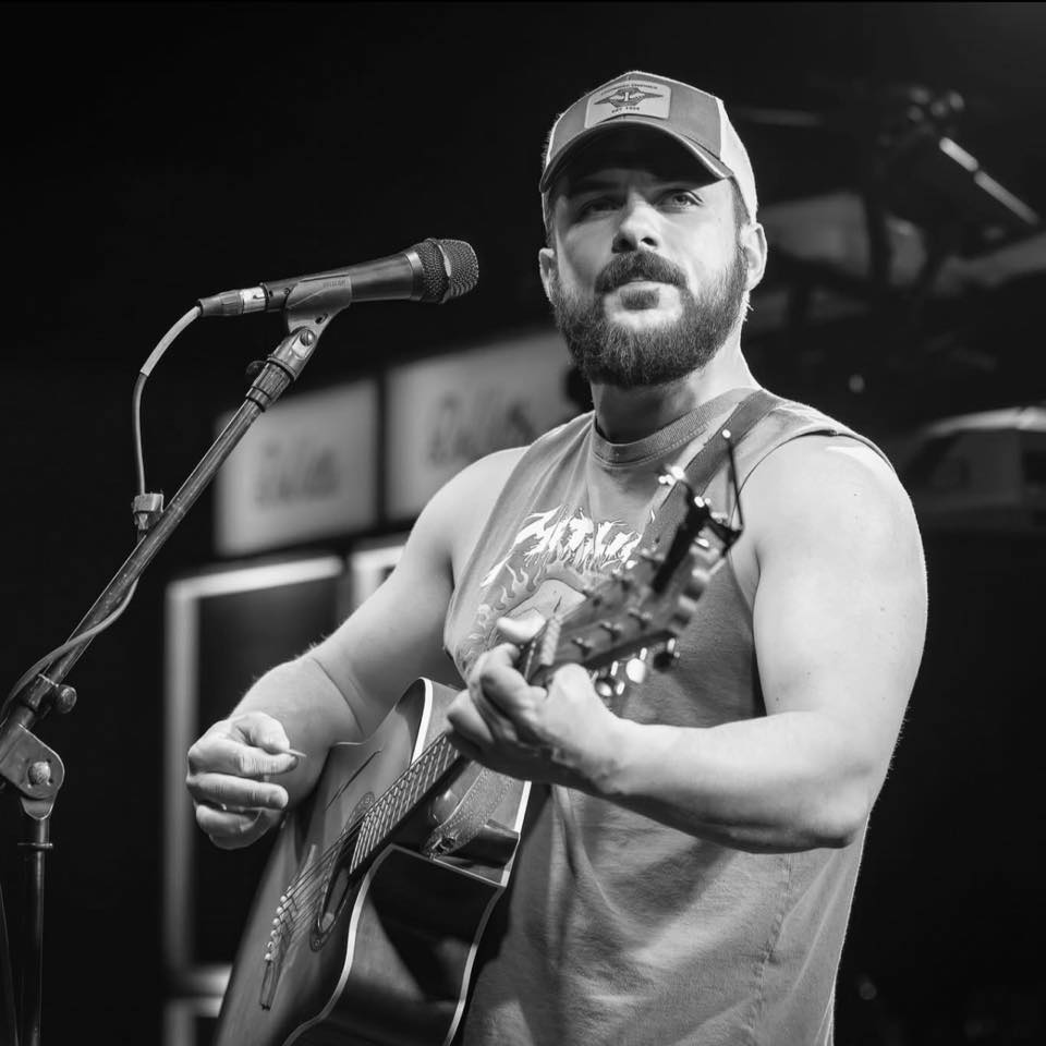 A man with a beard and a baseball cap plays an acoustic guitar and sings into a microphone.